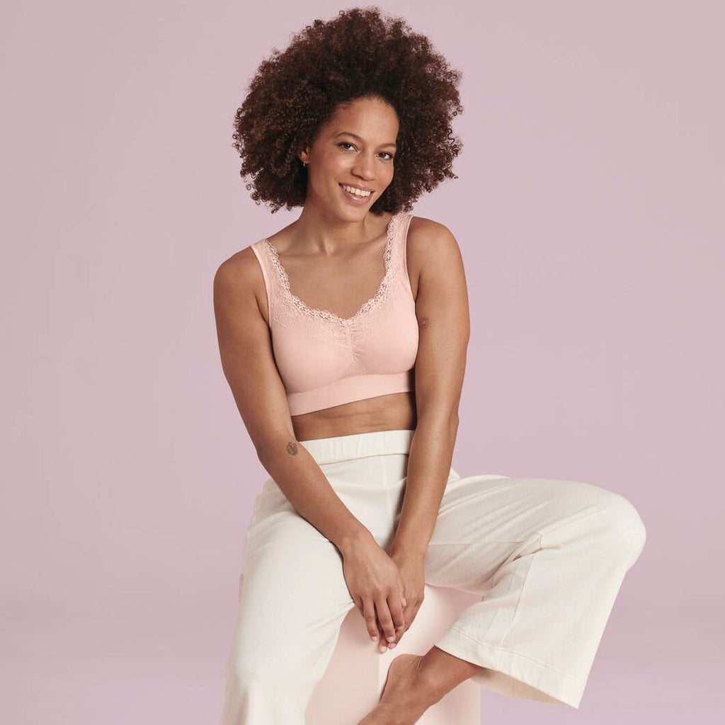 Woman sitting on a box wearing a pink bralette and white pants against a pink background
