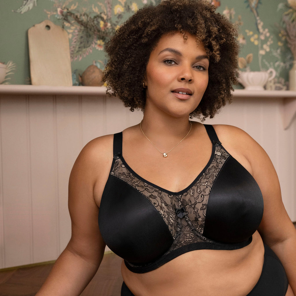 Woman wearing a black lace bra and underwear set in a room with floral wallpaper.